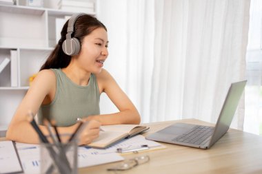 Beautiful young asian woman watching live video or video call of teacher teaching on laptop in her home, Take notes of important conversations and messages during the teacher's teaching.