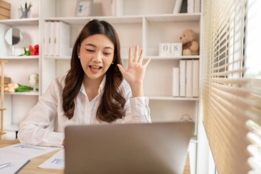 Genç Asyalı kadın, yüzünde gülümseme, internet iletişimi, bilgisayar, sosyal mesafe, internet öğrenimi ile arkadaşlarını veya meslektaşlarını canlı yayında selamlıyor..