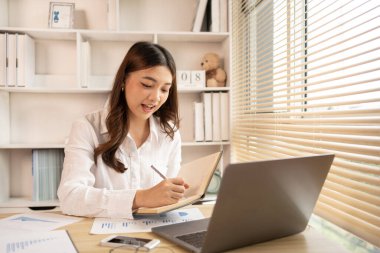 Beautiful young asian woman watching live video or video call of teacher teaching on laptop in her home, Take notes of important conversations and messages during the teacher's teaching.