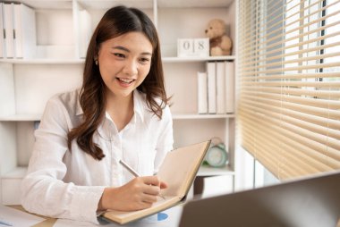 Beautiful young asian woman watching live video or video call of teacher teaching on laptop in her home, Take notes of important conversations and messages during the teacher's teaching.