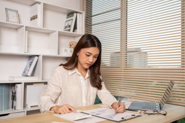 Evde çalışan Asyalı kadın, evin gelirini hesaplamak için hesap makinesine basın - evin giderlerini hesaplamak ve bilgiyi kaydetmek ve doğruluğunu kontrol etmek için dizüstü bilgisayarı kullanın..