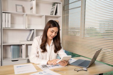 Evde çalışan Asyalı kadın, evin gelirini hesaplamak için hesap makinesine basın - evin giderlerini hesaplamak ve bilgiyi kaydetmek ve doğruluğunu kontrol etmek için dizüstü bilgisayarı kullanın..