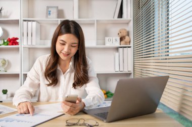 Asyalı kadın çalışan telefonda konuşuyor ve laptopta gülümseyen yüzüyle çalışıyor, ofis çalışanları çalışıyor ve telefon konuşmaları, hatırlatıcı notlar alıyor, telefonda iş konuşmaları yapıyor..