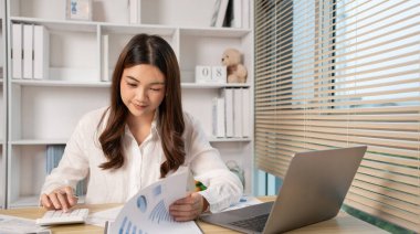 Evde çalışan Asyalı kadın, evin gelirini hesaplamak için hesap makinesine basın - evin giderlerini hesaplamak ve bilgiyi kaydetmek ve doğruluğunu kontrol etmek için dizüstü bilgisayarı kullanın..