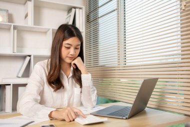 Evde çalışan Asyalı kadın, evin gelirini hesaplamak için hesap makinesine basın - evin giderlerini hesaplamak ve bilgiyi kaydetmek ve doğruluğunu kontrol etmek için dizüstü bilgisayarı kullanın..