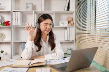 Genç Asyalı kadın, yüzünde gülümseme, internet iletişimi, bilgisayar, sosyal mesafe, internet öğrenimi ile arkadaşlarını veya meslektaşlarını canlı yayında selamlıyor..
