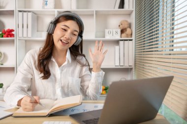 Beautiful young asian woman watching live video or video call of teacher teaching on laptop in her home, Take notes of important conversations and messages during the teacher's teaching.