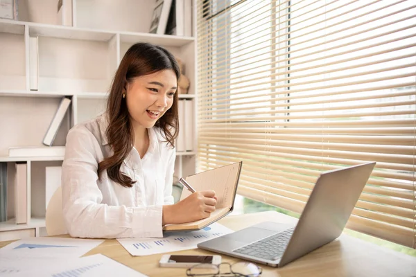 stock image Beautiful young asian woman watching live video or video call of teacher teaching on laptop in her home, Take notes of important conversations and messages during the teacher's teaching.