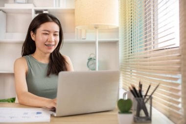 Woman using laptop to work or do homework at home with smiling face in her office, Creating happiness at work with a smile, Live performance or vdo call with laptop, Work from home.