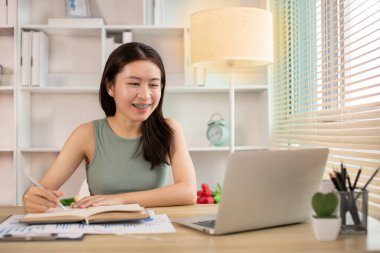 Beautiful young asian woman watching live video or video call of teacher teaching on laptop in her home, Take notes of important conversations and messages during the teacher's teaching.