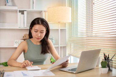 Evde çalışan Asyalı kadın, evin gelirini hesaplamak için hesap makinesine basın - evin giderlerini hesaplamak ve bilgiyi kaydetmek ve doğruluğunu kontrol etmek için dizüstü bilgisayarı kullanın..