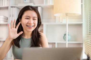 Genç Asyalı kadın, yüzünde gülümseme, internet iletişimi, bilgisayar, sosyal mesafe, internet öğrenimi ile arkadaşlarını veya meslektaşlarını canlı yayında selamlıyor..