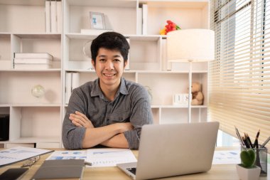 Yakışıklı genç adam, elinde laptop ve ofis ekipmanıyla masa başında oturuyor ve hafta başında çalışmaya hazır gülümsüyor. Üretken Çalışma Haftasına Pozitif Başlangıç.