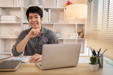 Yakışıklı genç adam, elinde laptop ve ofis ekipmanıyla masa başında oturuyor ve hafta başında çalışmaya hazır gülümsüyor. Üretken Çalışma Haftasına Pozitif Başlangıç.