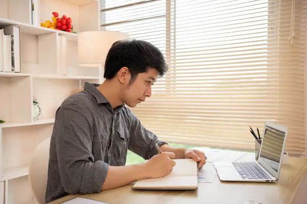 Studying online using a laptop, Man watching live video or video call of teacher teaching on laptop in her home, Take notes of important conversations and messages during the teacher's teaching.
