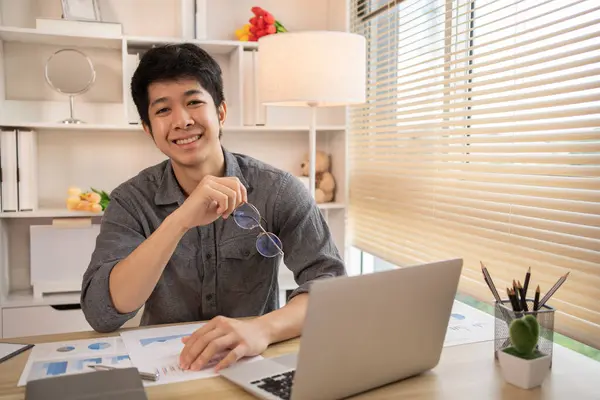 Yakışıklı genç adam, elinde laptop ve ofis ekipmanıyla masa başında oturuyor ve hafta başında çalışmaya hazır gülümsüyor. Üretken Çalışma Haftasına Pozitif Başlangıç.