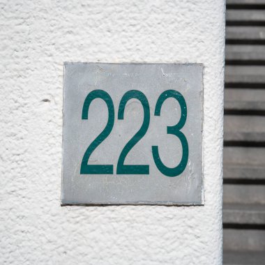 Beyaz alçılı bir duvara monte edilmiş metal bir plaka üzerinde '223' (223) yazan yeşil metal bir ev numarası..