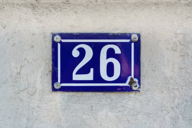 '26' (26) yazan hasarlı bir maden ev numarası, beyaz alçılı bir duvara sabitlenmiş bir plakette..