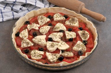 Domatesli mozzarella turtası, merdane ve gri arka planda çay havlusuyla pişirmeden önce bir tabakta hazırlandı.