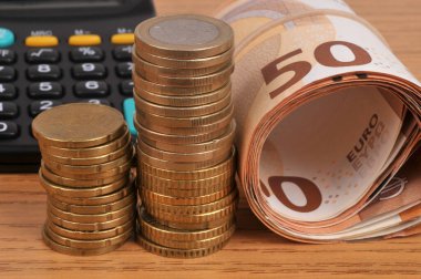 Stacks of euro coins with rolled up banknotes and a calculator in the background clipart