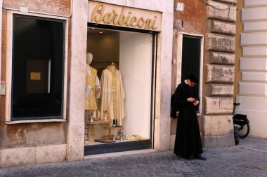 Roma 'da bir caddedeki elbise ve dini eşyalar dükkanının yanındaki akıllı telefonuyla rahip.