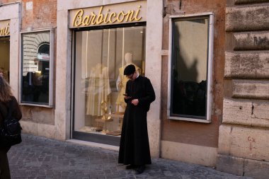 Roma 'da bir caddedeki elbise ve dini eşyalar dükkanının yanındaki akıllı telefonuyla rahip.