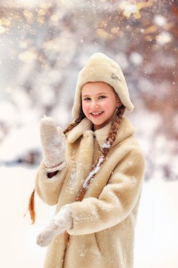 Beyaz örülü şapkalı neşeli küçük bir kız, çam ağacının sessiz kış ormanında karlı bir yolda duruyor ve büyük bir soğuk kartopu tutuyor.