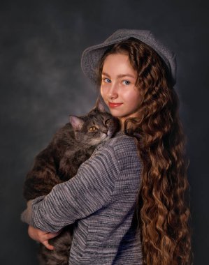 Uzun kıvırcık saçlı, eskimiş elbiseli, gülümseyen kız stüdyo resminde gri kediyi tutuyor. Kedi şakayla dudaklarını yalıyor.