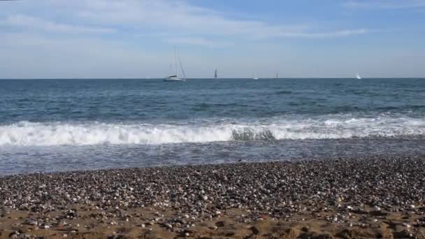 地中海のきれいなビーチ 砂や小石の上に海の波 地平線にヨット 海岸に白い海泡が形成されます 海の休暇 ビーチリゾート 夏の時間 海の上のヨット 海岸沿いの散歩 — ストック動画
