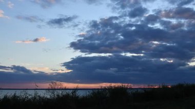 Gün batımında bulutların dingin göl suyunun üzerinde geniş gökyüzünde sürüklenmesiyle günün geceye geçişi. Renkler çarpıcı bir şekilde değişir, ufku aydınlatır.