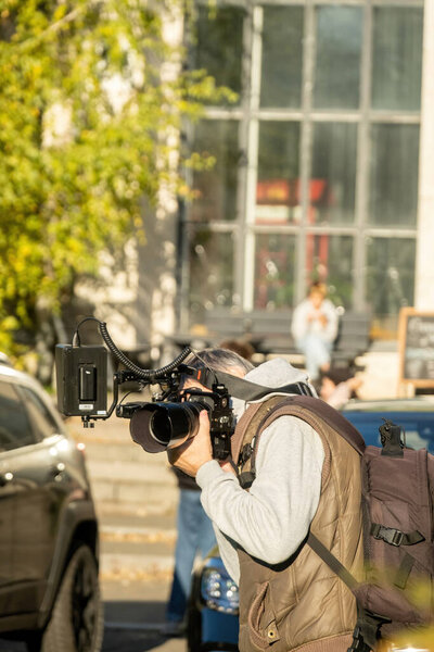 Adult male photographer stands on driveway near cars with professional camera closing face. Grey-haired man with backpack in sportswear shoots on spring day