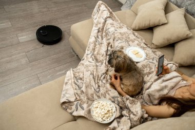 Battaniyeli kadınla kaplı bej koltukta akıllı telefona bakıyor. Shih Tzu, dükkan sahibinin yanında oturmuş çalışan robotlu elektrik süpürgesine bakıyor.