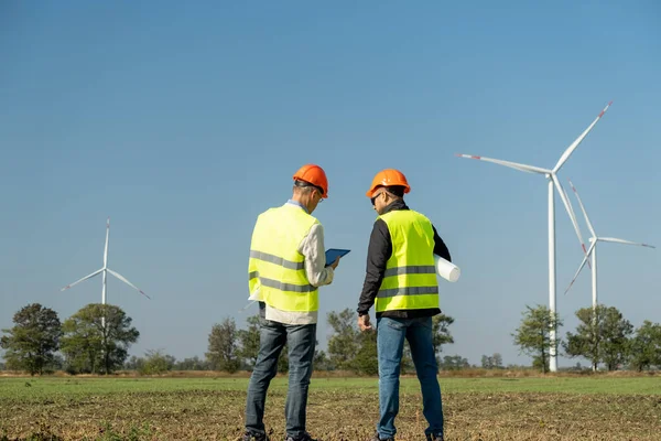 風車は田舎で代替エネルギーを生産する ヘルメットのメンテナンスの技術者は 風力発電機に対するプロジェクトについて話しますバックビュー — ストック写真