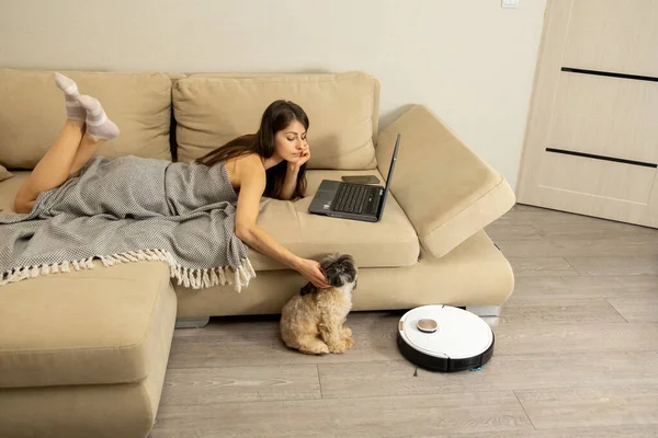 Dog sits near sofa and white robotic vacuum cleaner. Covered with blanket long-haired woman lies on sofa with laptop and pets Shih Tzu in light apartment