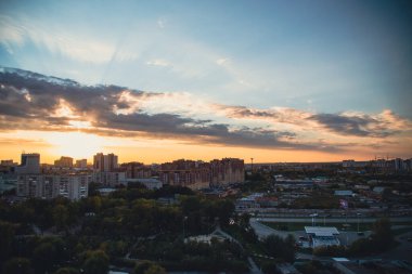 Gün ışığı bulutların arasından doğar ve akşam şehrini aydınlatır. Yol üzerinde çok katlı binalar yükseliyor. Arabalar karanlık ağaçlarla çevrili.
