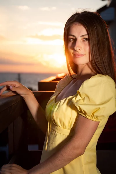 Young beautiful woman sits sideways holding on wooden railing of balcony. Dark-haired lady looks straight spending vacation alone in hotel at seaside