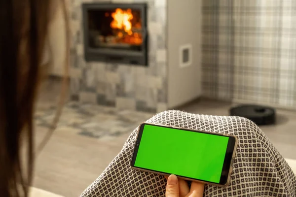 Woman looks at mobile telephone screen with chromakey. Person sits in cozy room with fireplace and robotic vacuum cleaner on blurred background closeup