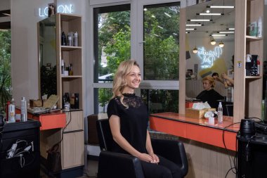 KYIV, UKRAINE - 27 EPTEMBER 2021: Siyah bluzlu sarışın kadın büyük aynalı makyaj sanatçısının işyerinin yanındaki sandalyede oturuyor. Modern güzellik salonunda mutlu bir gülümseme.