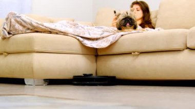 Robot elektrikli süpürge oturma odasının zeminini temizliyor. Kadın cep telefonuyla postaları kontrol ediyor. Köpek arkadaşıyla evdeki kanepede yatıyor.