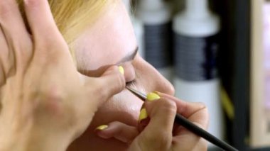 Kadın usta, sarışın müşteri için doğal makyaj yapıyor. Sanatçının manikürlü elleri güzellik salonunun arka planında göz kapağına kozmetik fırçayla siyah ok çizer.