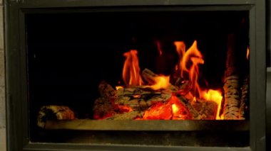 Modern şöminede yanan parlak ateş oturma odasına sıcaklık saçıyor. Alevlerin turuncu dilleri yakın görüşte yanmış odunlarda parıldıyor.