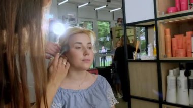 KYIV, UKRAINE - 27 Eylül 2021: Profesyonel kadın sanatçı sarışın müşterisiyle güzellik salonunda çalışıyor. Esmer kadın makyaj yapıyor. Yanaklarına ve alnına allık sürüyor.