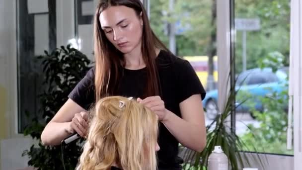 懸念ブルネットの美容師は 顧客の長いブロンドの髪にカール鉄を使用しています 女性のクライアントは 美容室でパーティーのために準備アームチェアに座っている — ストック動画