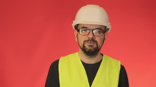 戴着白色头盔的专业建筑工人在镜头前看着 工作室特写中 大胡子专家靠红墙站立的肖像 — 图库视频影像