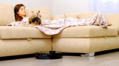 Robot süpürgesi oturma odasındaki katları temizliyor. Telefonu olan genç bir kadın ve tüylü bir köpek arkadaşı evdeki kanepede dinleniyor.