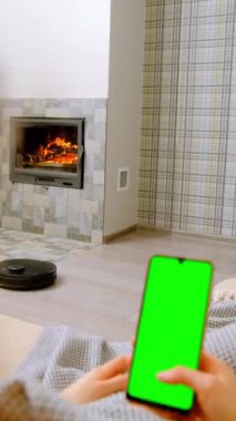 Robot elektrikli süpürge oturma odasındaki şöminenin yanındaki lamine zeminde çalışır. Kadınlar kanepeye uzanır. Yakın görüşte akıllı telefon kullanır.