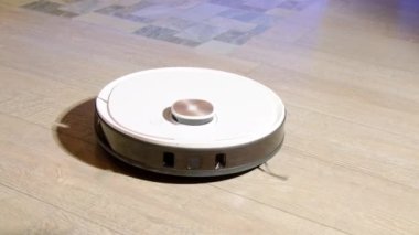 Beyaz robot süpürgesi oturma odasındaki lamine ahşap zemindeki tozu temizliyor. Modern cihazlar ev işlerine yardımcı olur. Yerli aygıt yakın görünüm