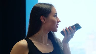 Genç bir kadın sesli mesaj dinler ve pencerenin yanında gülümser. Sarkık küpeli kadın telefonu tutuyor ve dinlenme önermesine gülümsüyor.