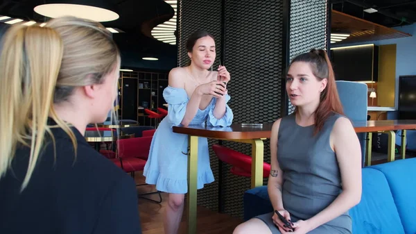 Young women talk and relax during break at resting premise. Woman in blue dress enjoys leisure and drinks coffee with straw at coworking lounge