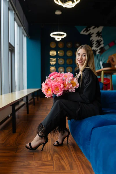 Blonde woman sits with flower arrangement on couch in staff lounge room. Female employee holds birthday gift with contented expression